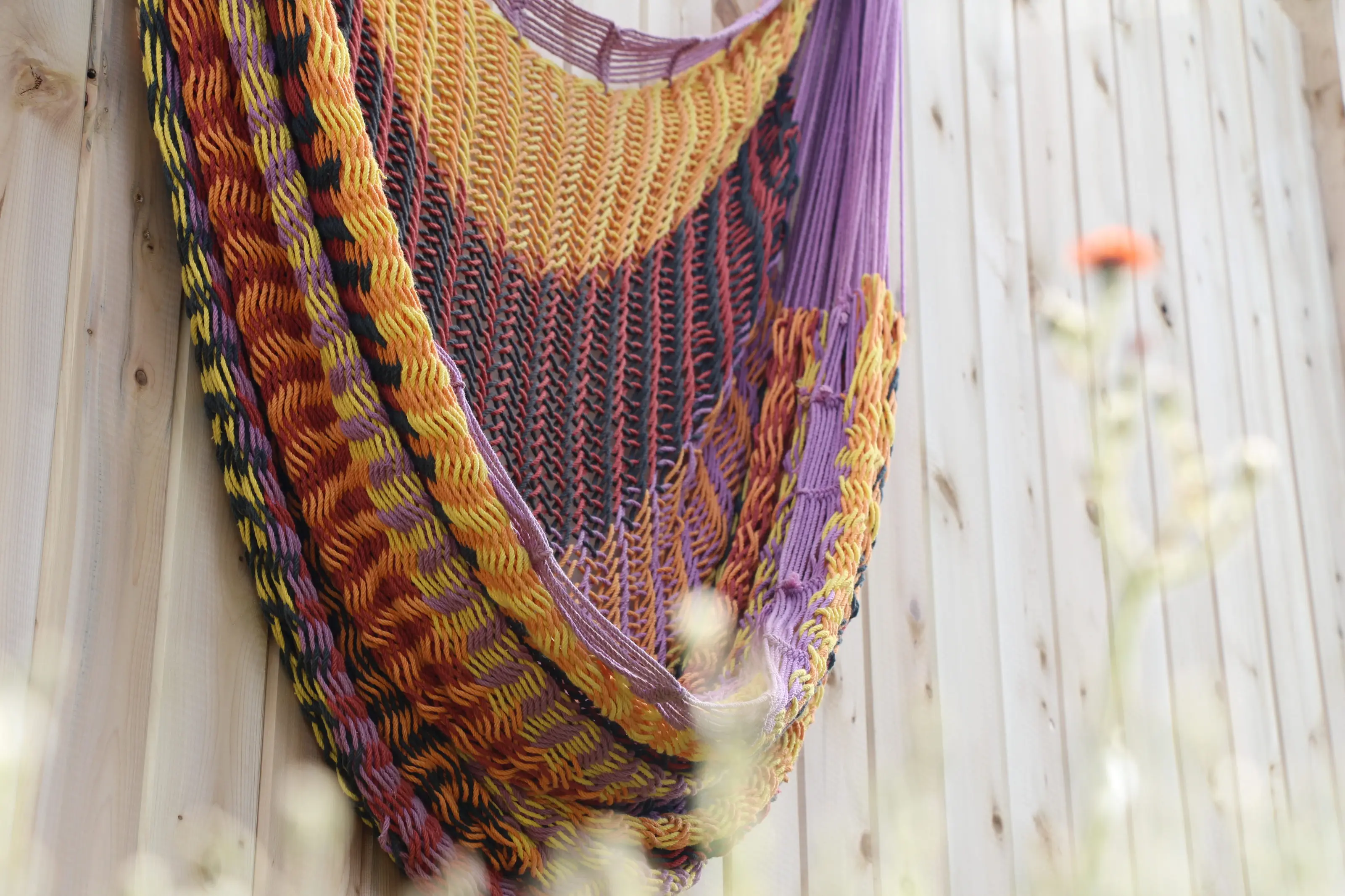 Un hamac tissé de terracotta et de violet, suspendu contre un mur de cèdre, des fleurs sauvages au premier plan